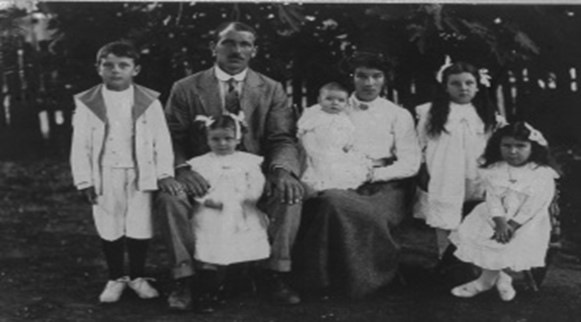 Image: Old black and white photograph of a large family, man and woman with five young children, sitting in the garden posing for the photographer.