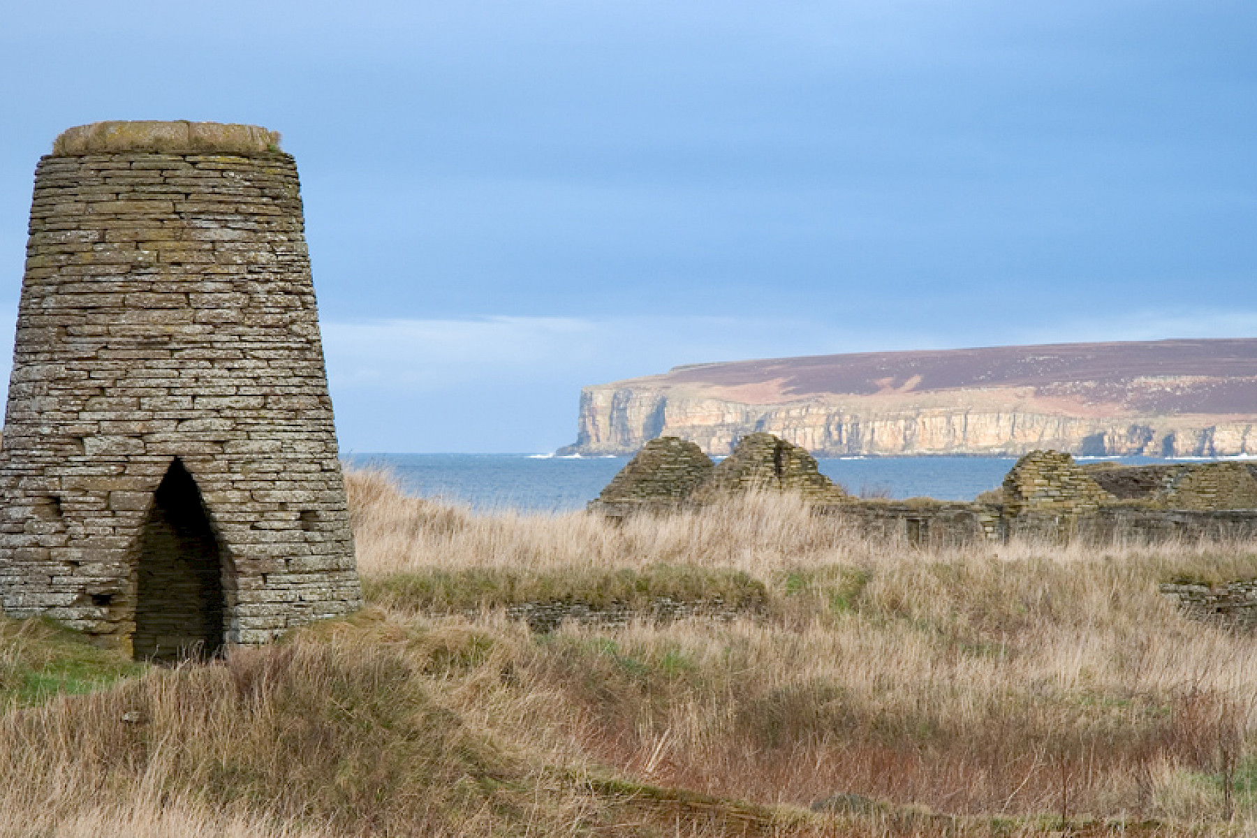 Castletown image: Stone tower monument with a small door stands on an open cliff top with distant headland and blue sea in the background.