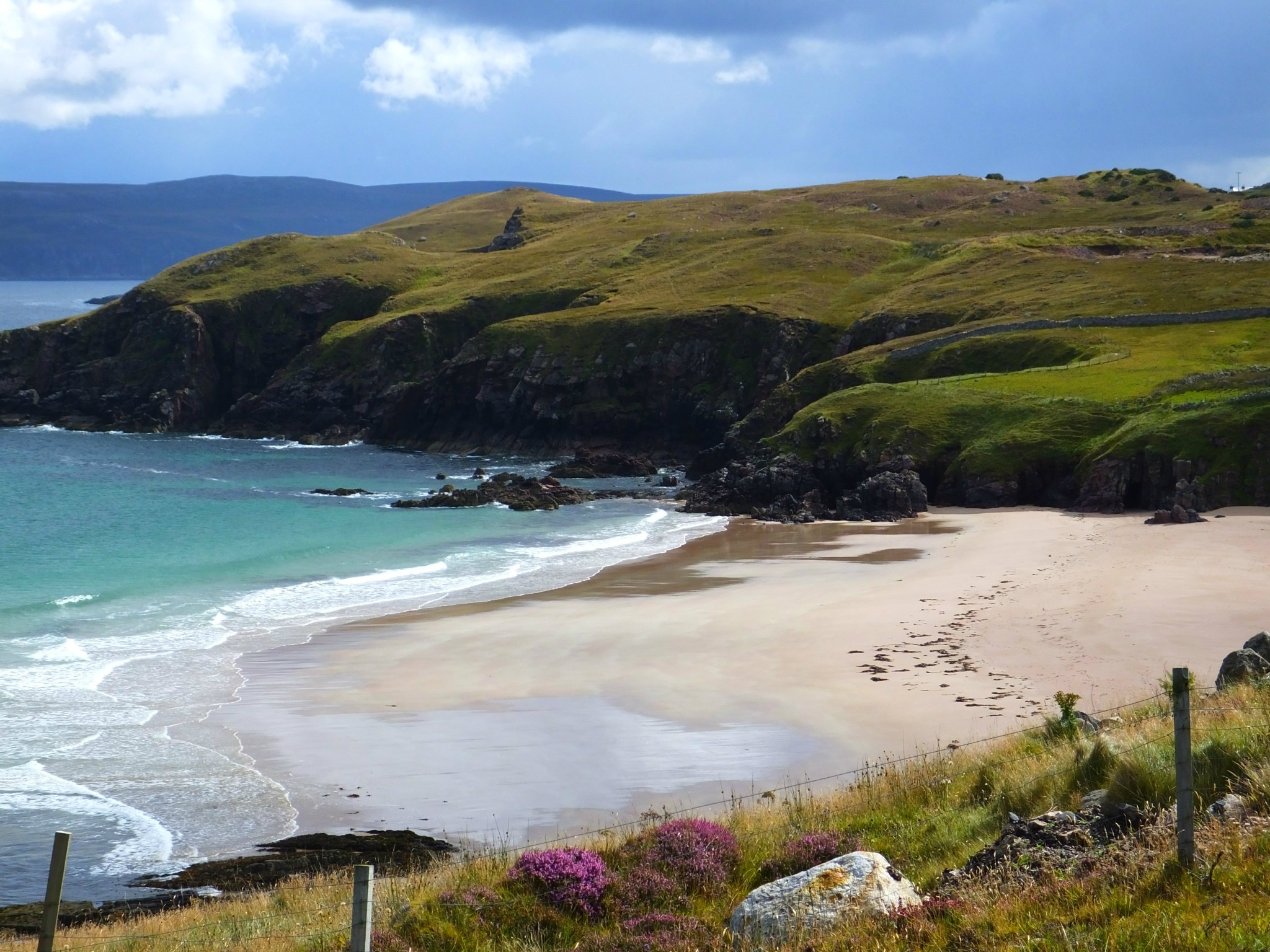 Durness Beach - Golden sands lie in front of a gentle green headland and white waves wash up on the beach.