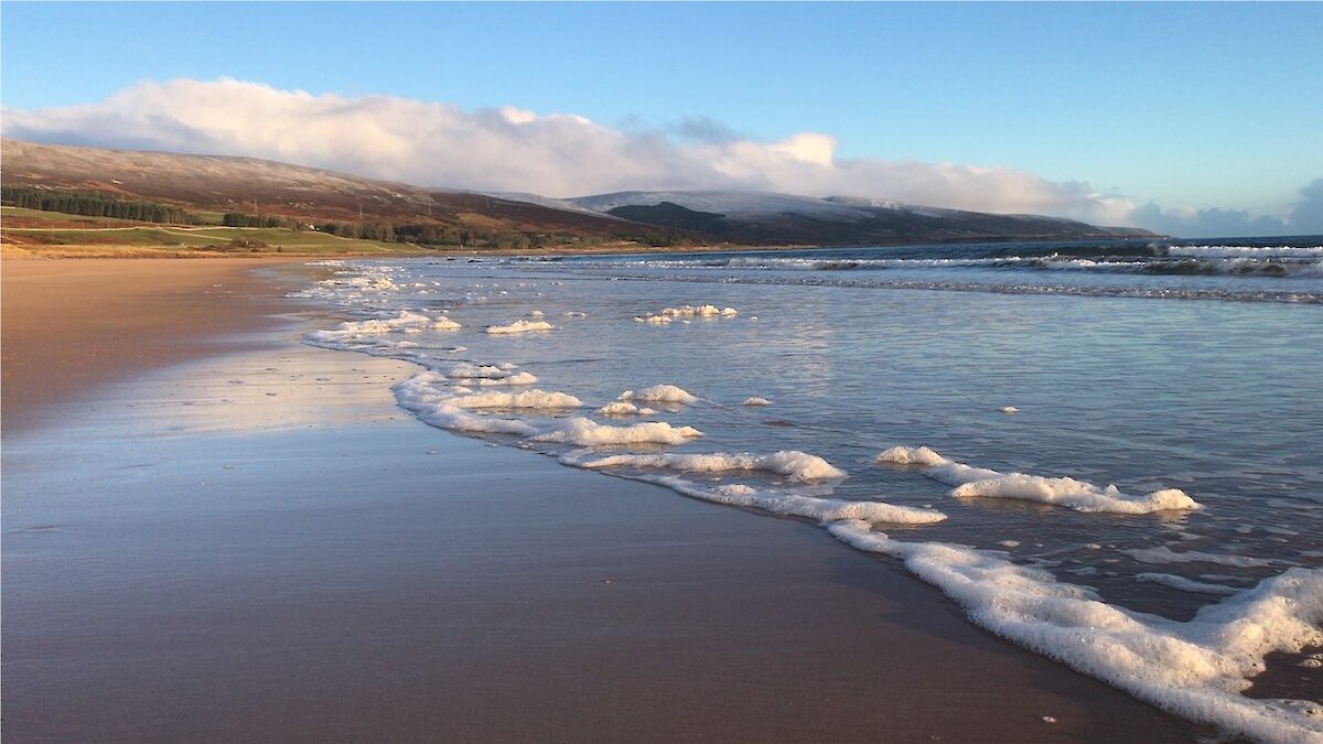 Wide sandy beach is washed with gentle drifts of sea spray while snow covered hills mark the horizon.