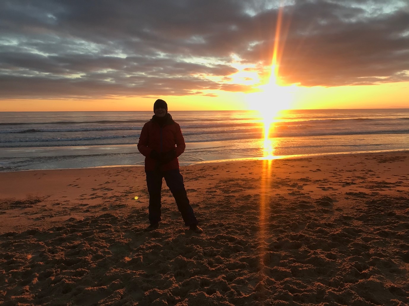 Amanda stands silhouetted by a fiery sunset on a sandy beach with gentle ocean waves in the background.