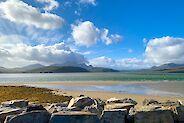 Bright blue sky with puffy white clouds look down over a stunning vista of a wide beach with playful water behind and some far off rugged mountains in the background.