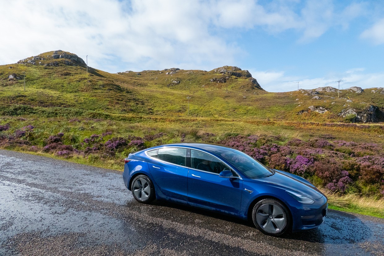 Dark blue electric car parks up on a sloped single track road with rocky hills covered in bright purple heather and rough undergrowth behind.