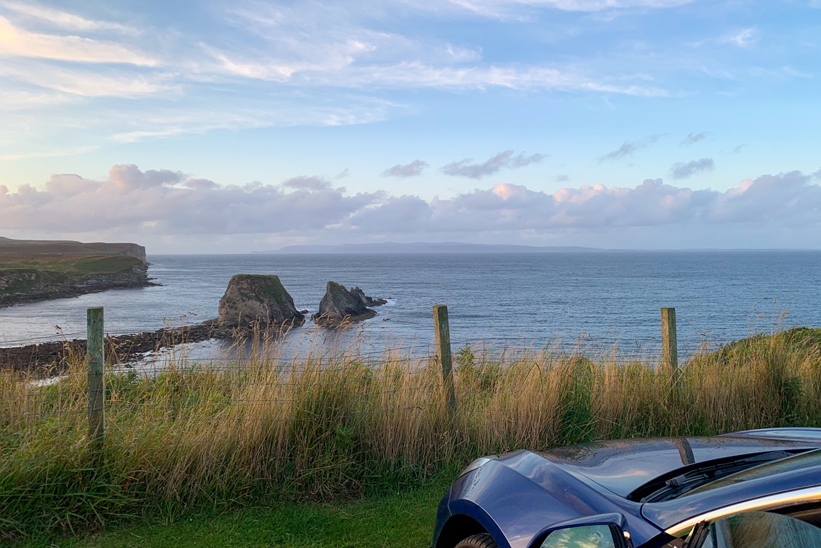 Front of a car faces out to the horizon where a wide open vista of sea, cliffs and jagged rocks lie beyond a simple wire fence. A far off island can be seen in the distance.