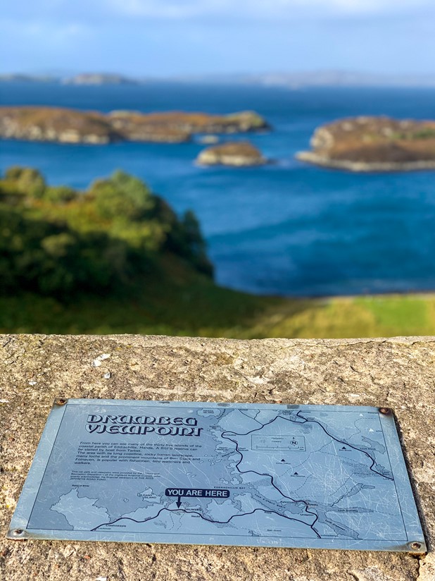 Drumbeg Viewpoint, shows a map of the area on a stone plinth while an out-of-focus background shows crystal blue waters dotted with small rocky islets.