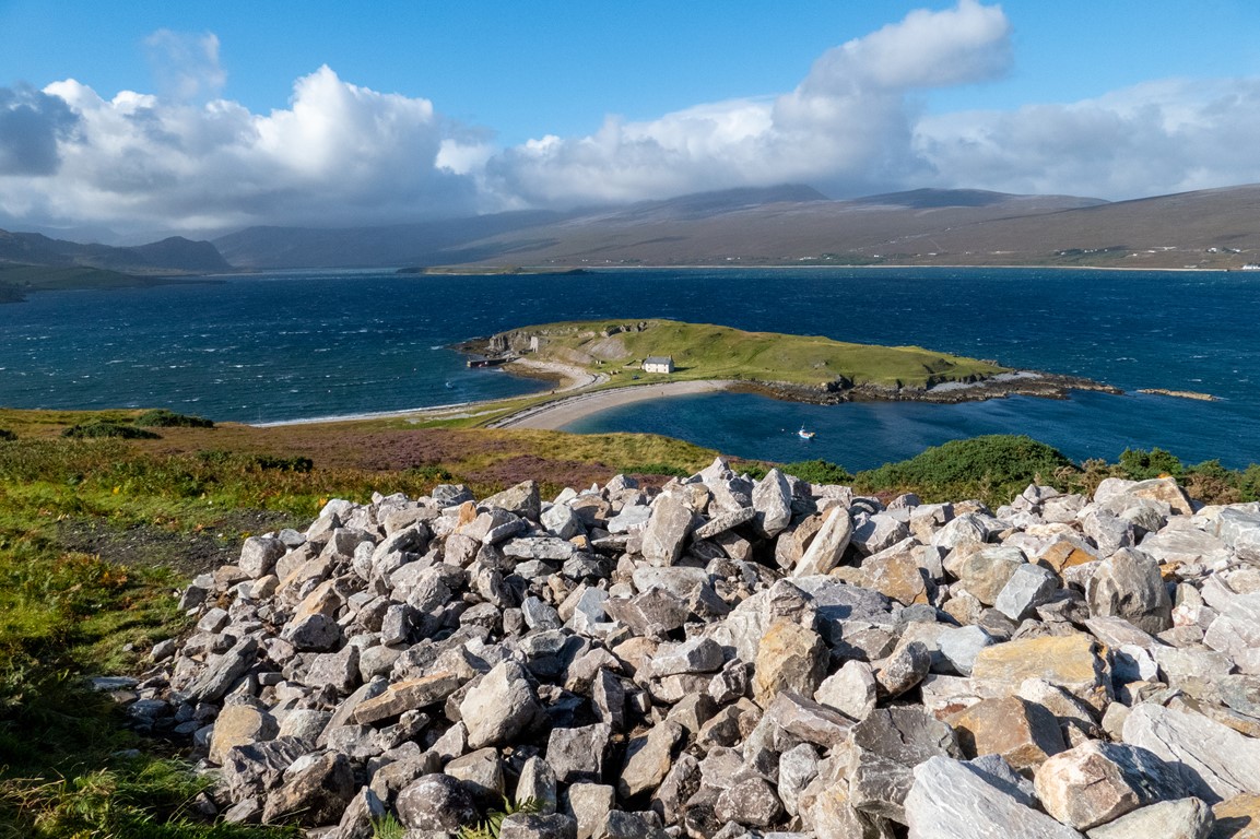 A small white cottage sits far below surrounded on all sides by deep blue waters and only a small sandy causeway connecting its grassy island to the mainland.