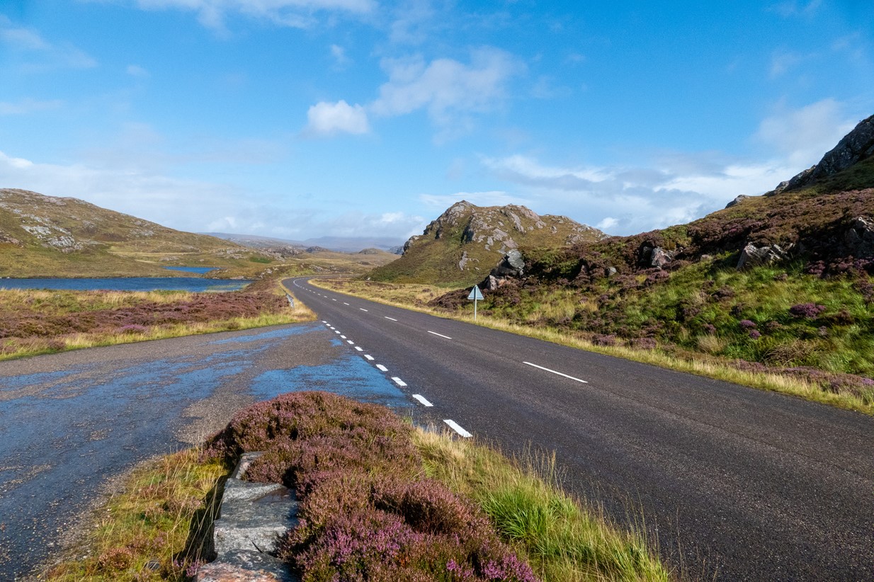 A wide open road stretches into the distance before disappearing round a bend with rocky hills surrounding it on both sides and the end of a loch just peeping into the horizon.