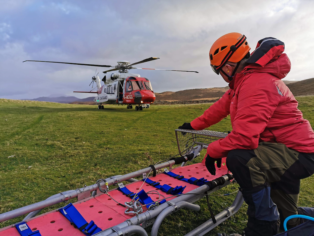 Image: Mountain Rescue volunteer crouches by an empty stretcher as a helicopter lands on the grass not too far away.