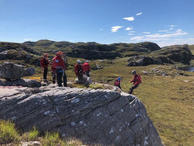 Image: Two mountain rescue volunteers practice lowering a stretcher down a rock face on a bright sunny day.