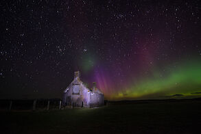 Image: Northern Lights flicker in the sky behind a ruined stone cottage.