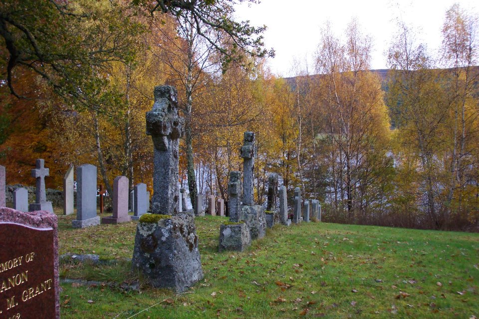 Image: Modern colour photo of celtic crosses in a graveyard surrounded by autumn trees with a loch behind.