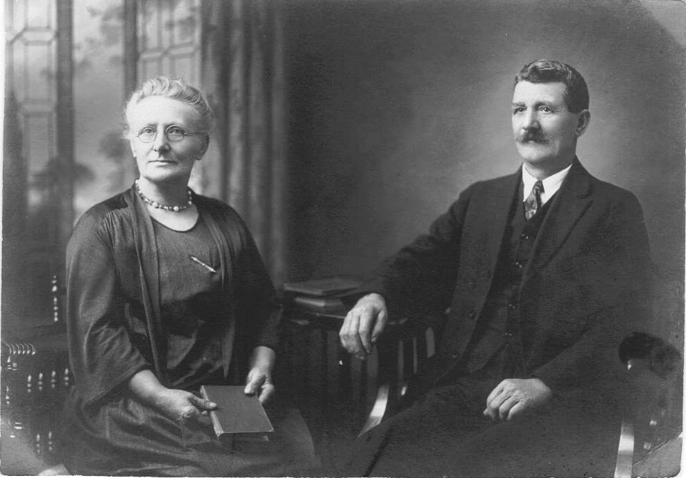 Image: Old black and white photograph of an older man and woman sitting in chairs posing for the photographer.