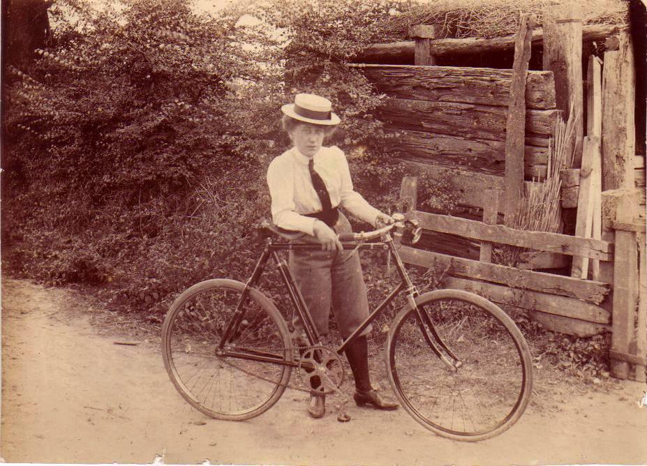 Image: Old black and white photograph of a young woman wearing a boater hat and holding on to an bicycle.