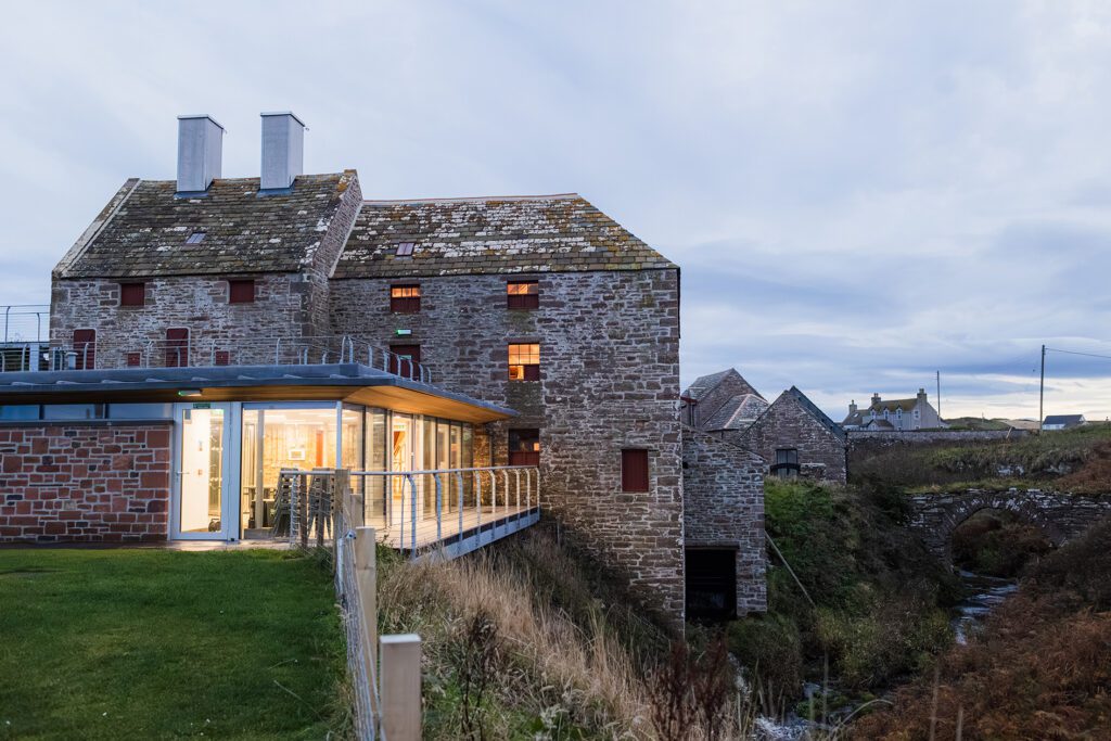 Early evening view of John O’Groats Mill with warm lights glowing inside, showcasing its cozy community space and heritage architecture.