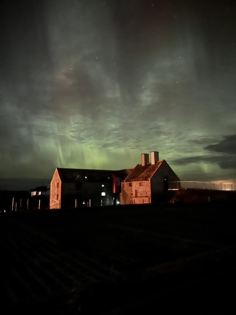 A historic stone watermill at John O’Groats illuminated by warm lights under a dramatic night sky filled with green and faint purple aurora borealis streaks.