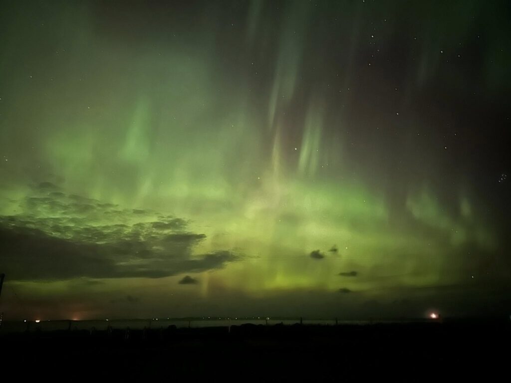 A sweeping view of the Caithness coastline at John O’Groats under a vivid green aurora borealis display.