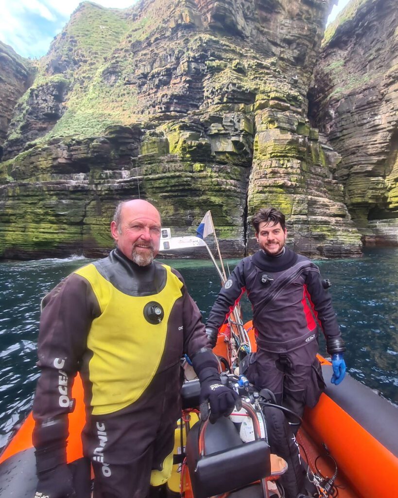 Two divers stood on a boat out at sea, a large cliff stands behind them. Activities and sport in the North Highlands