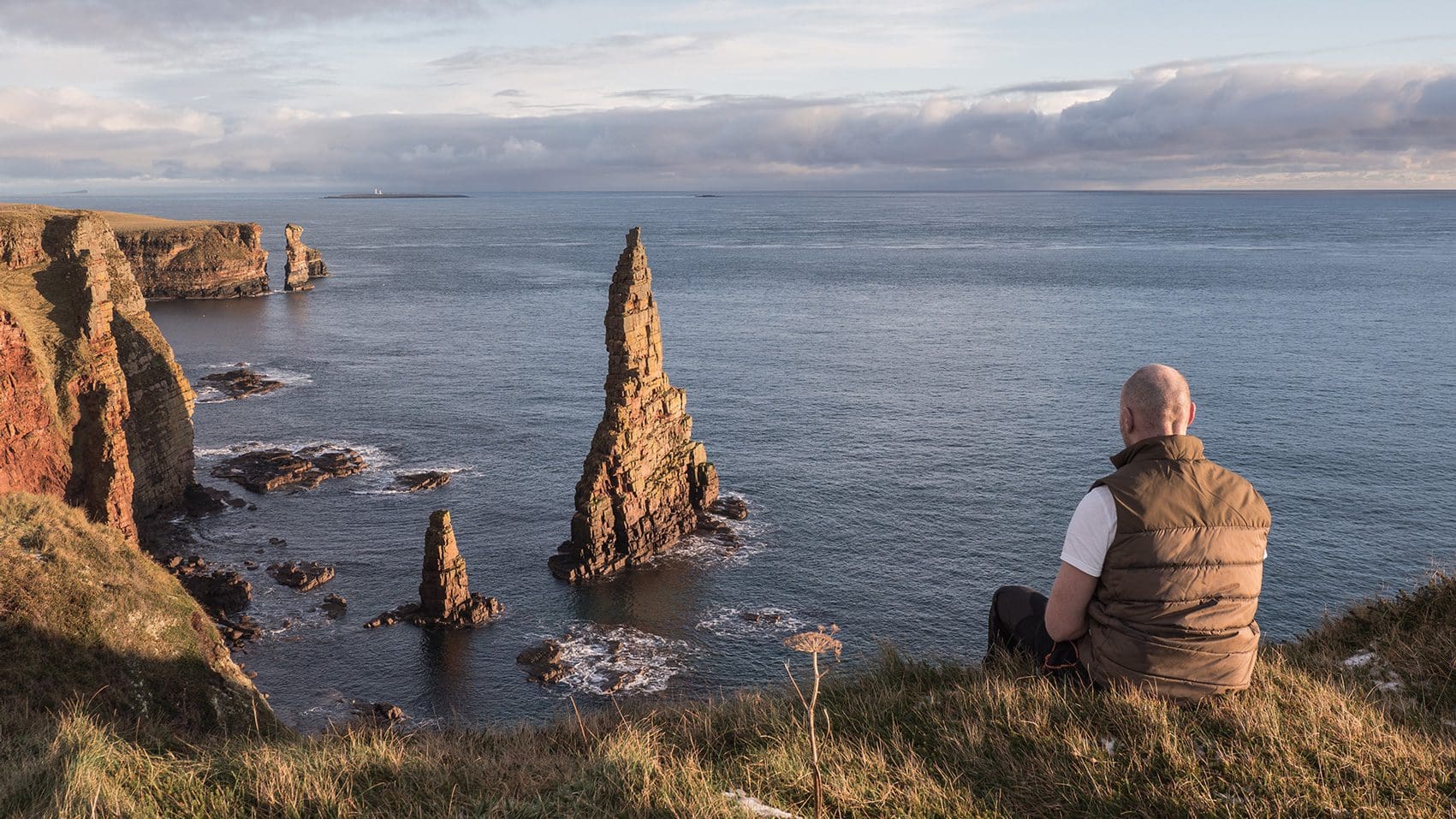 Person sitting on grassy cliff overlooking Duncansby Stacks sea stacks near John O’Groats, Scotland’s far north coast, with calm ocean and dramatic rock formations of the Duncansby Stacks.