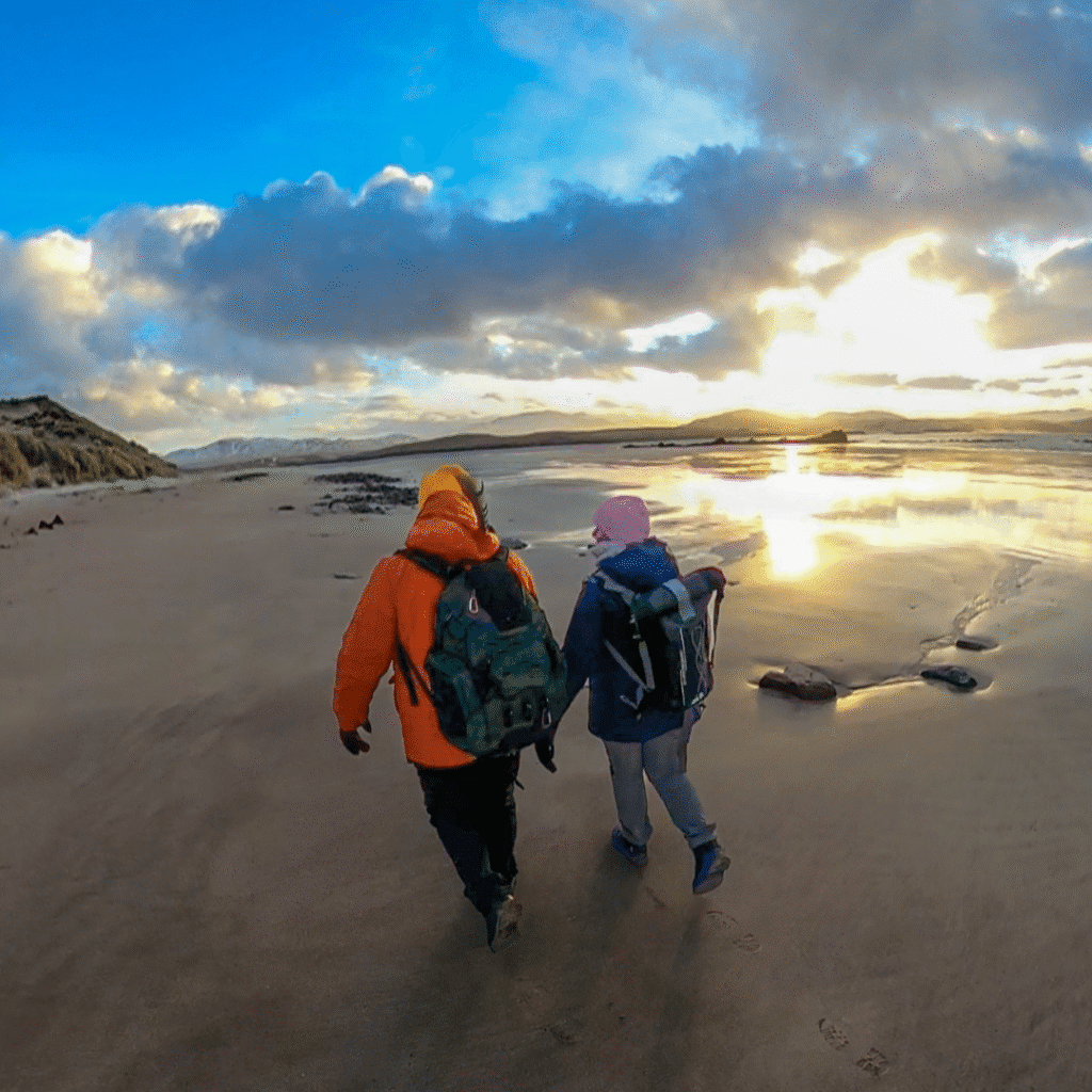 Two people walk on a beach, the sunsetting in the background. In Sutherland, Scotland.