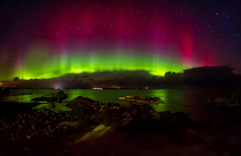 Aurora over Sinclair Bays water with greens and reds in the sky, in Caithness, Scotland.