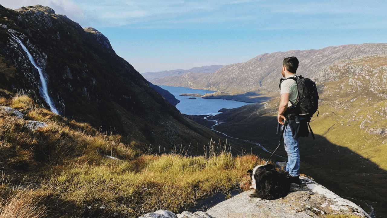 A man stands on a hill, overlooking hills and a lake in the distance. In Assynt, Sutherland, Scotland.