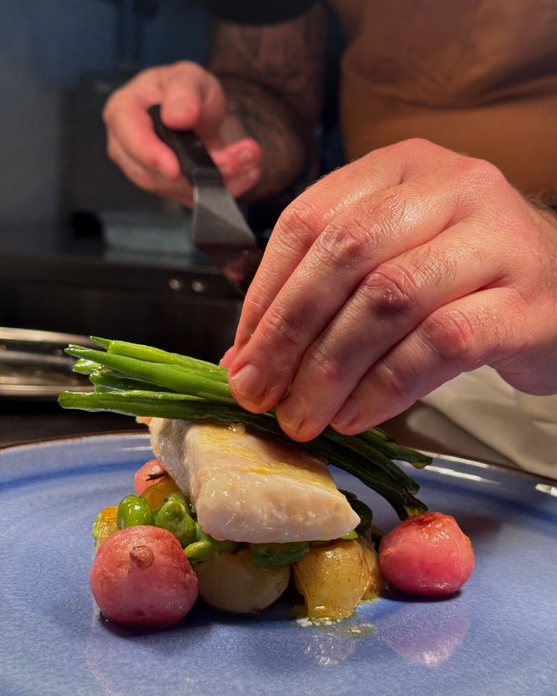In a restaurant in Scotland a man is putting together a plate, placing greenbeans ontop of a piece of Salmon.