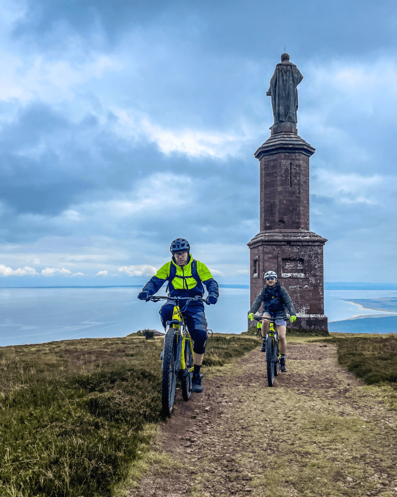 Two people cycling on Ben Bhraggie, in Golspie, Scotland. The statue of the 1st Duke of Sutherland in the background.