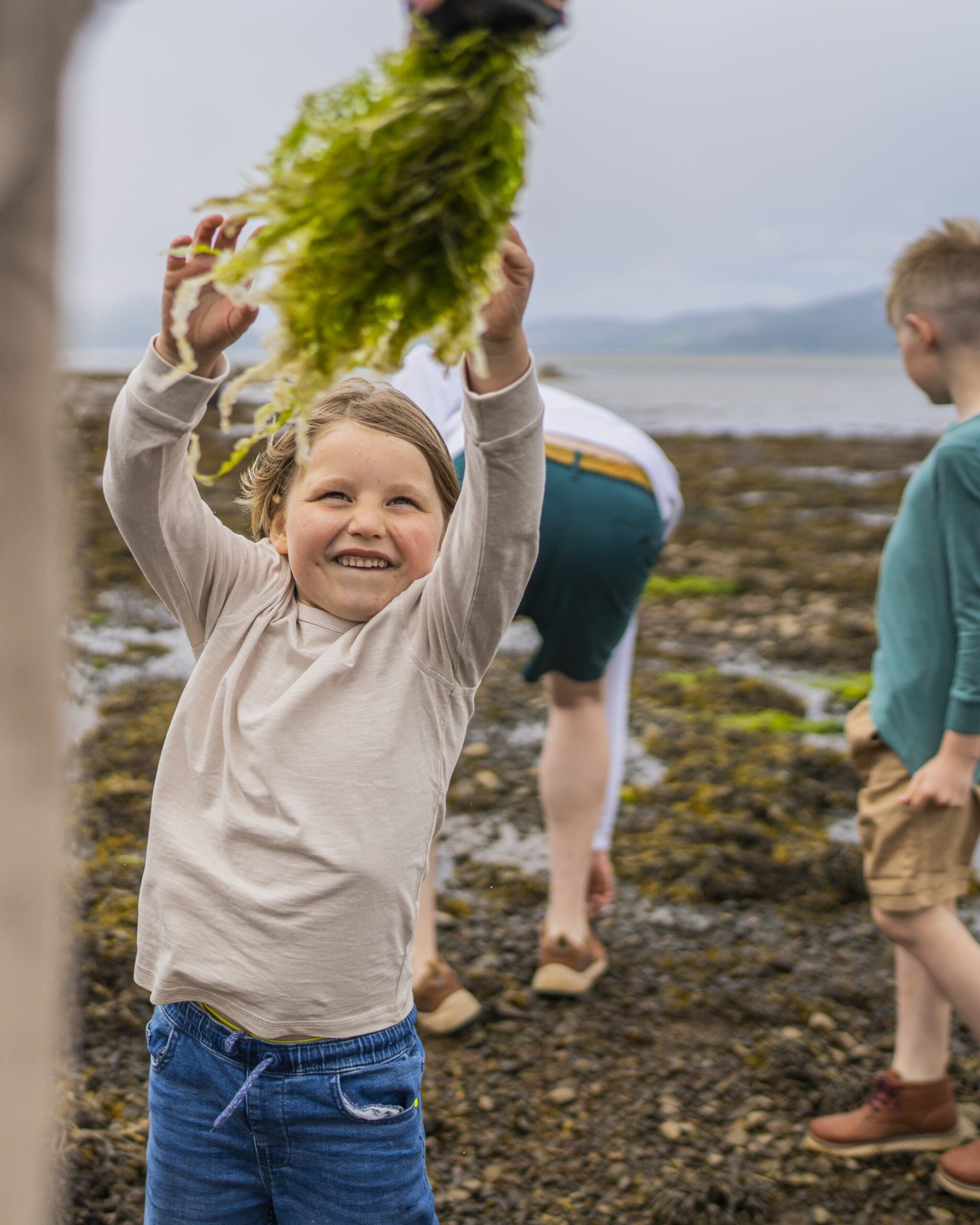 Children playing on the bank of Loch Fleet, the child in front is reaching for some seaweed, smiling. Sutherland, Scotland. nature and outdoors in the North Highlands