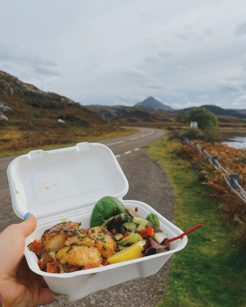 A person holding their takeaway food, a view of Loch Clash behind them, in Sutherland, Scotland.
