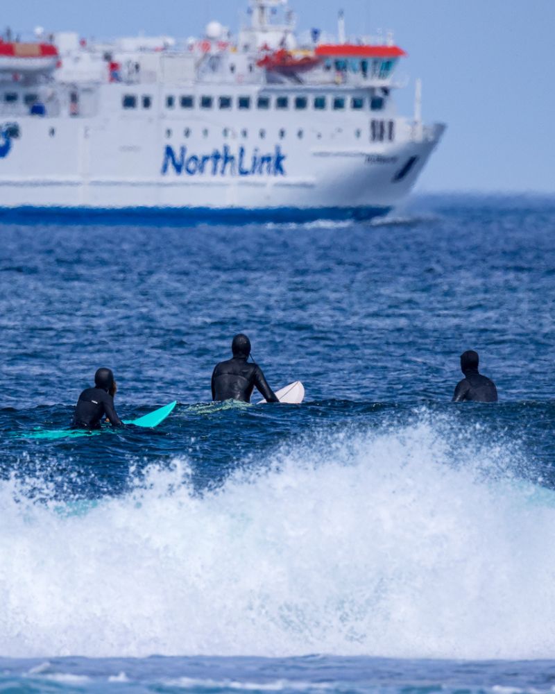Three surfers float on the waves, they are looking at a North Link ferry passing in the distance. Activities and sport in the North Highlands