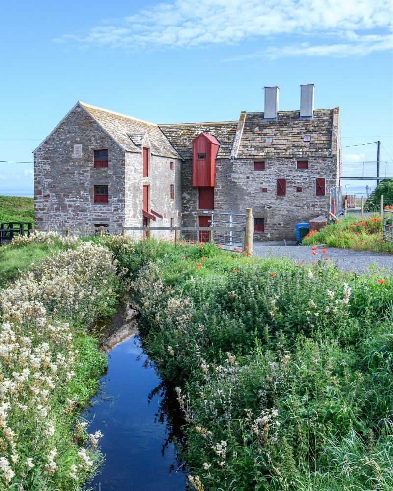 The John O'Groats Mill, in Caithness, Scotland, the building is viewed from the river naxt to it, with long green grass and clear blue sky. Attractions and landmarks.