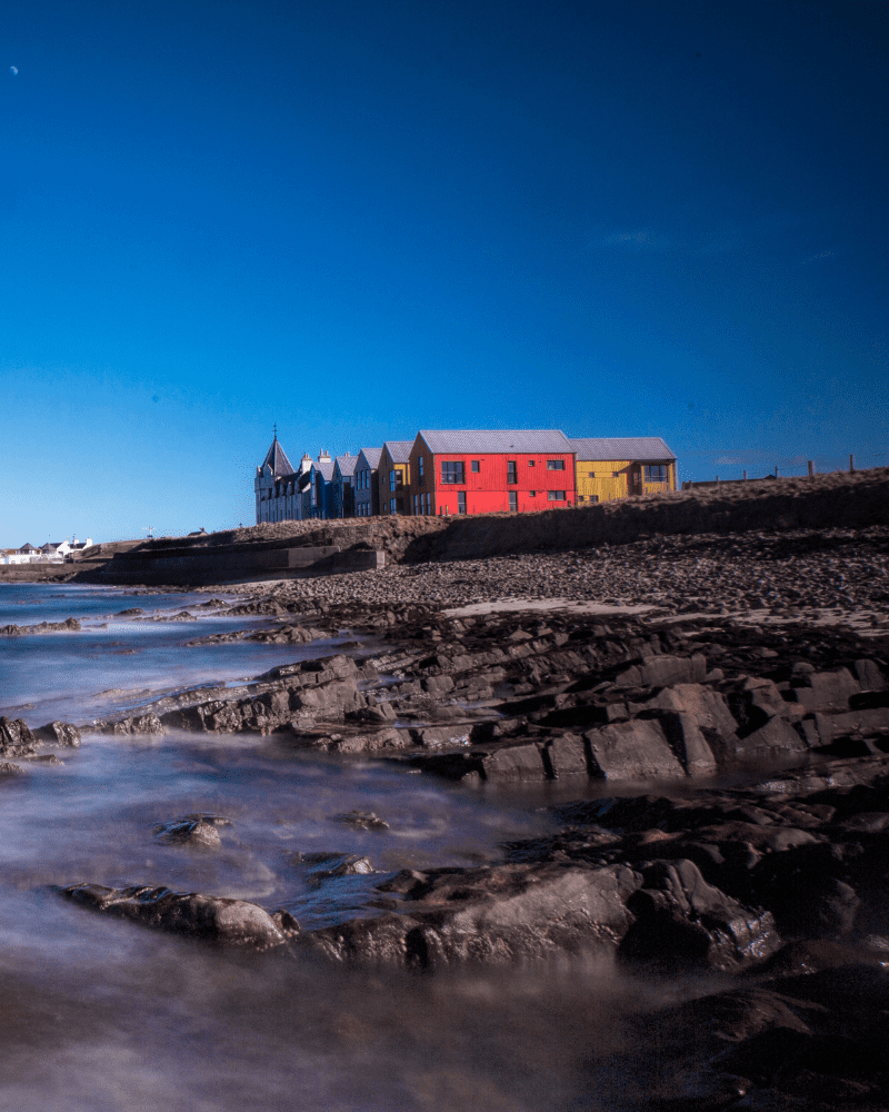 The Inn at John O'Groats, a array of colourful buildings viewed from the beach, with a clear blue sky. Attractions and landmarks.