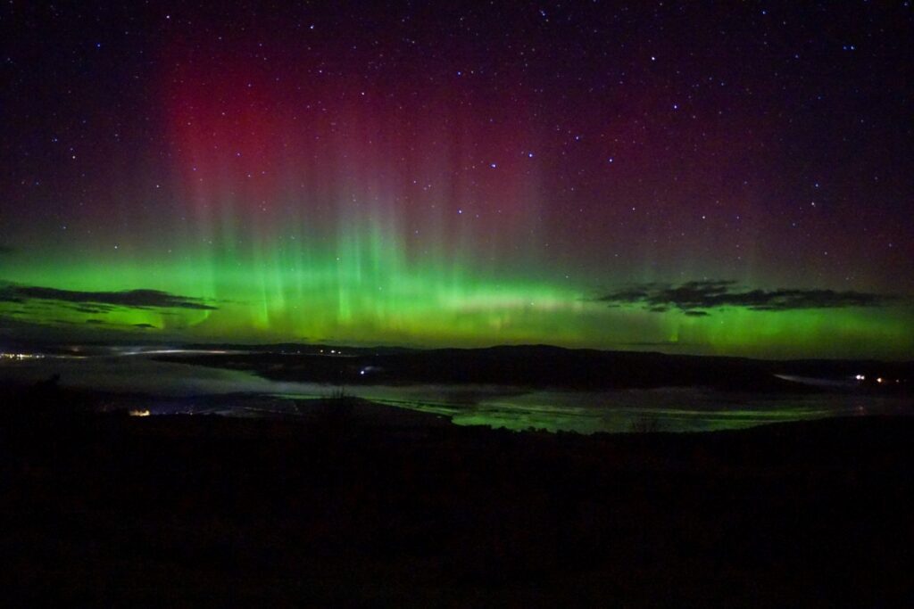 Northern Lights reflected on the water at the Kyle of Sutherland, with vibrant green and pink, purple aurora above a tranquil Highland landscape.