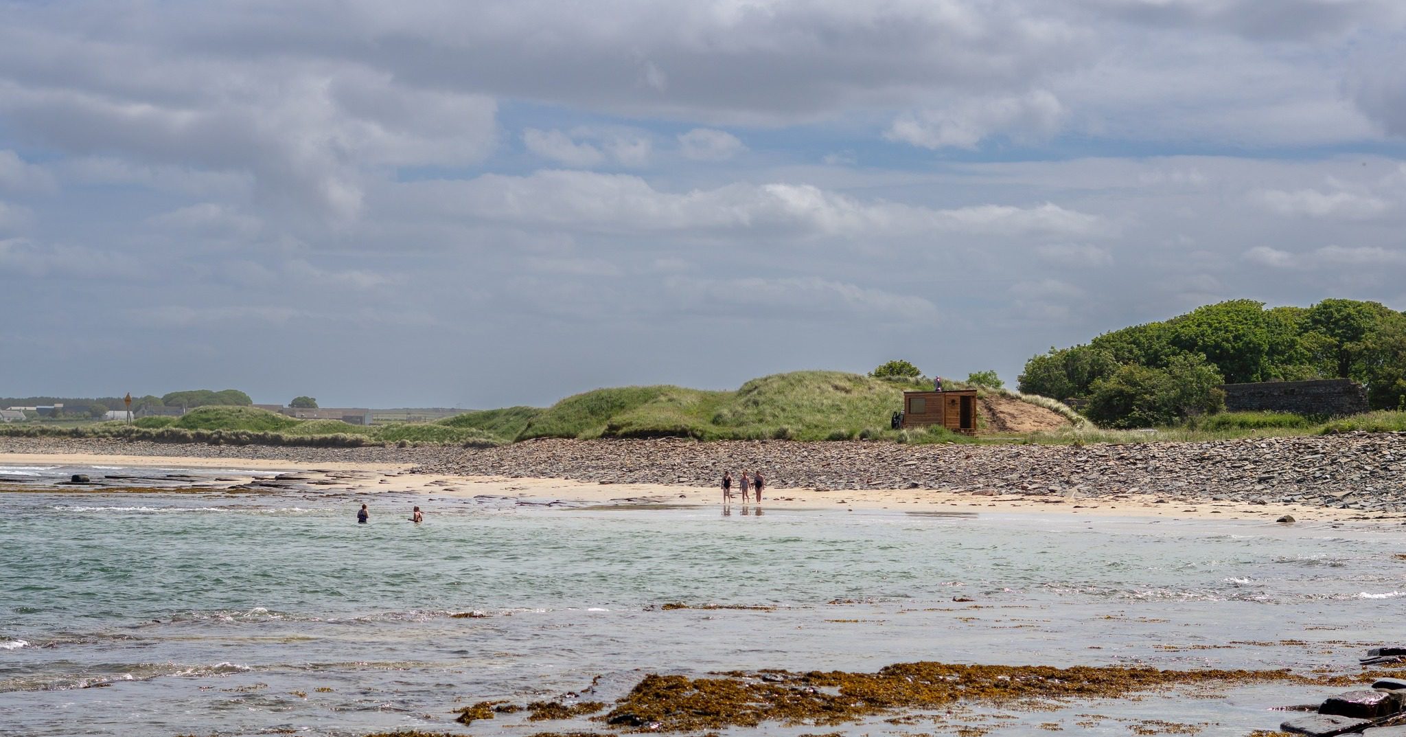 People wading in the sea with the Clach Mara Sauna on the sand behind them, at Castletown beach in Caithness, Scotland