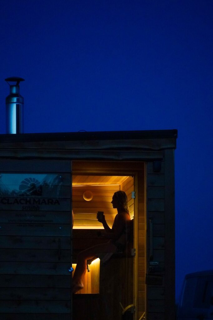 A woman sits inside the Clach Mara Sauna, holding a drink, at night, located in the North Highlands