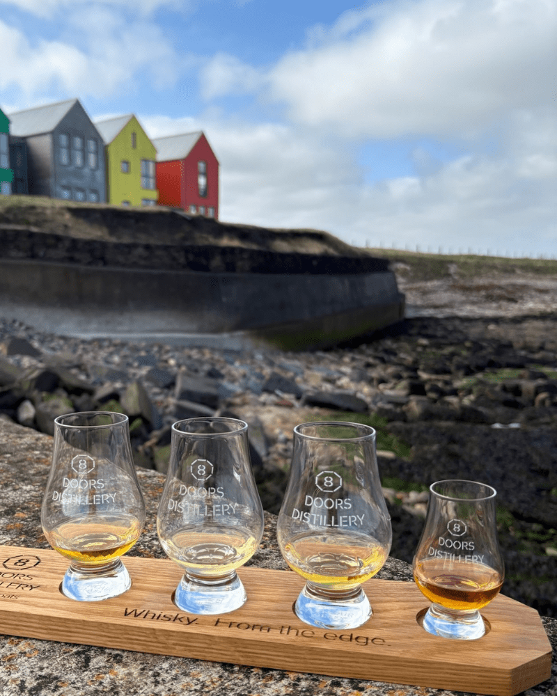 Four glasses of whiskey, with the 8 Doors Distillery logo, John O'Groats can bee seen in the background.