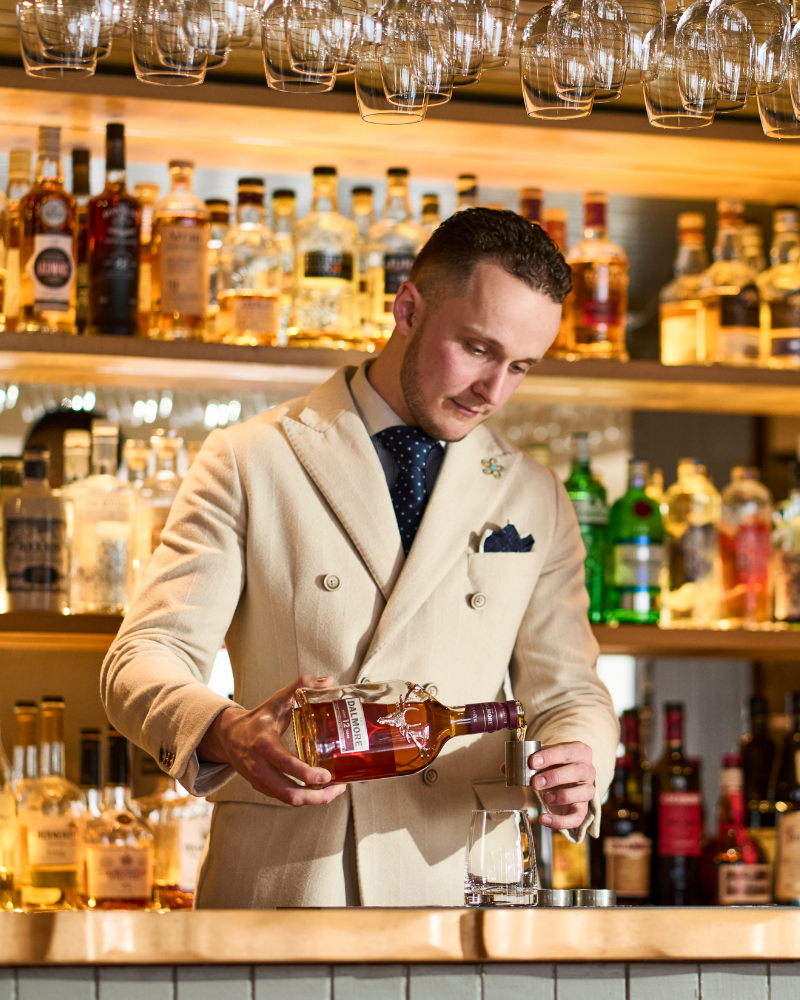 A bartender pouring a drink, with a golden background of drinks on shelves. Highland Coast Hotels, Kylesku, Sutherland, Scotland.