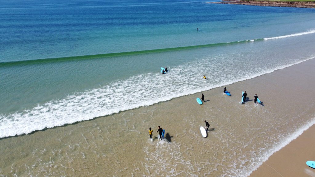 Surf lesson at Dunnet Beach on Scotland’s NC500, with beginners carrying surfboards into clear turquoise water under sunny skies.