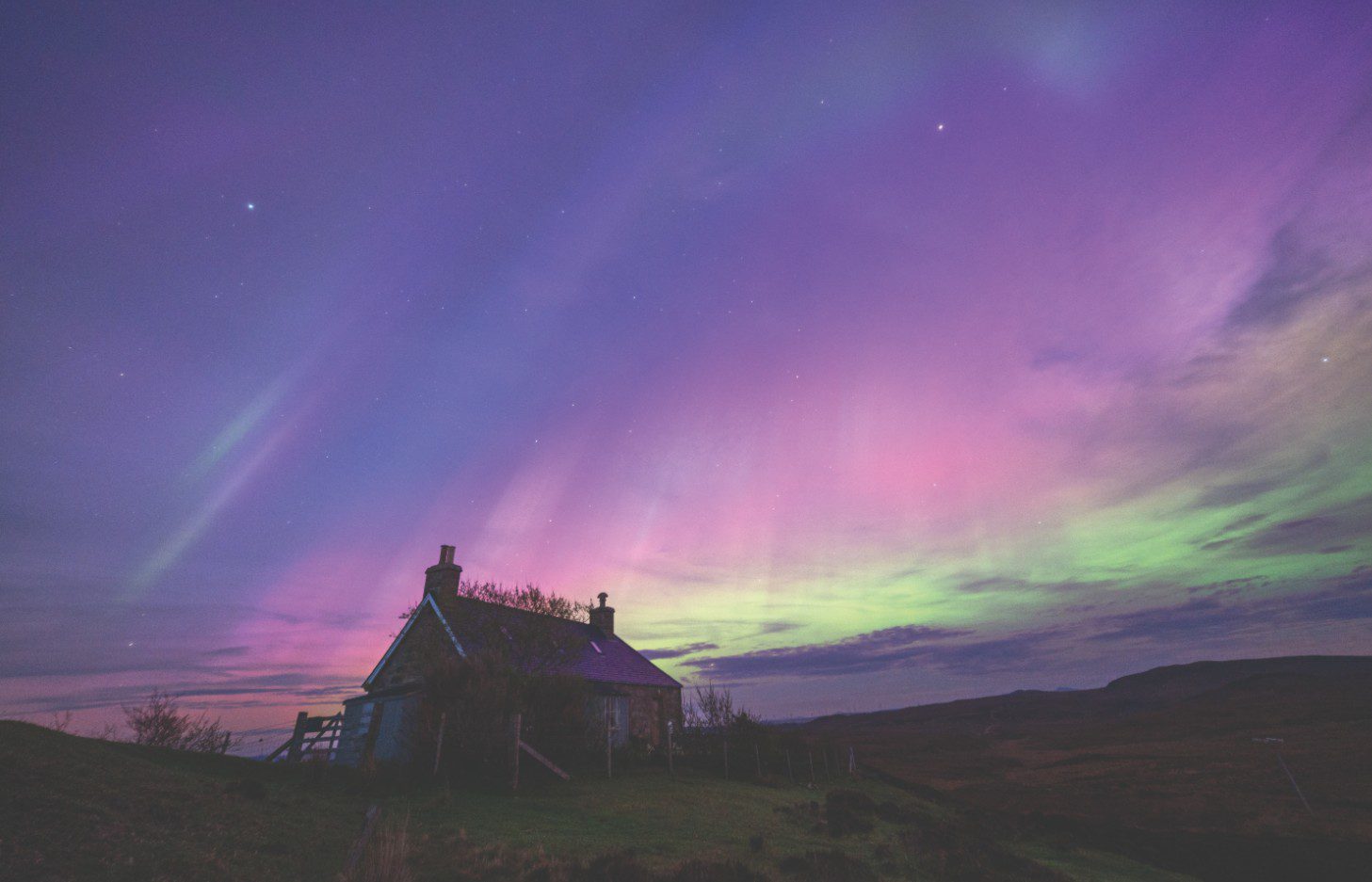 Northern Lights in vibrant purple, pink, and green hues illuminating the night sky above a rural cottage in Sutherland, Scotland.