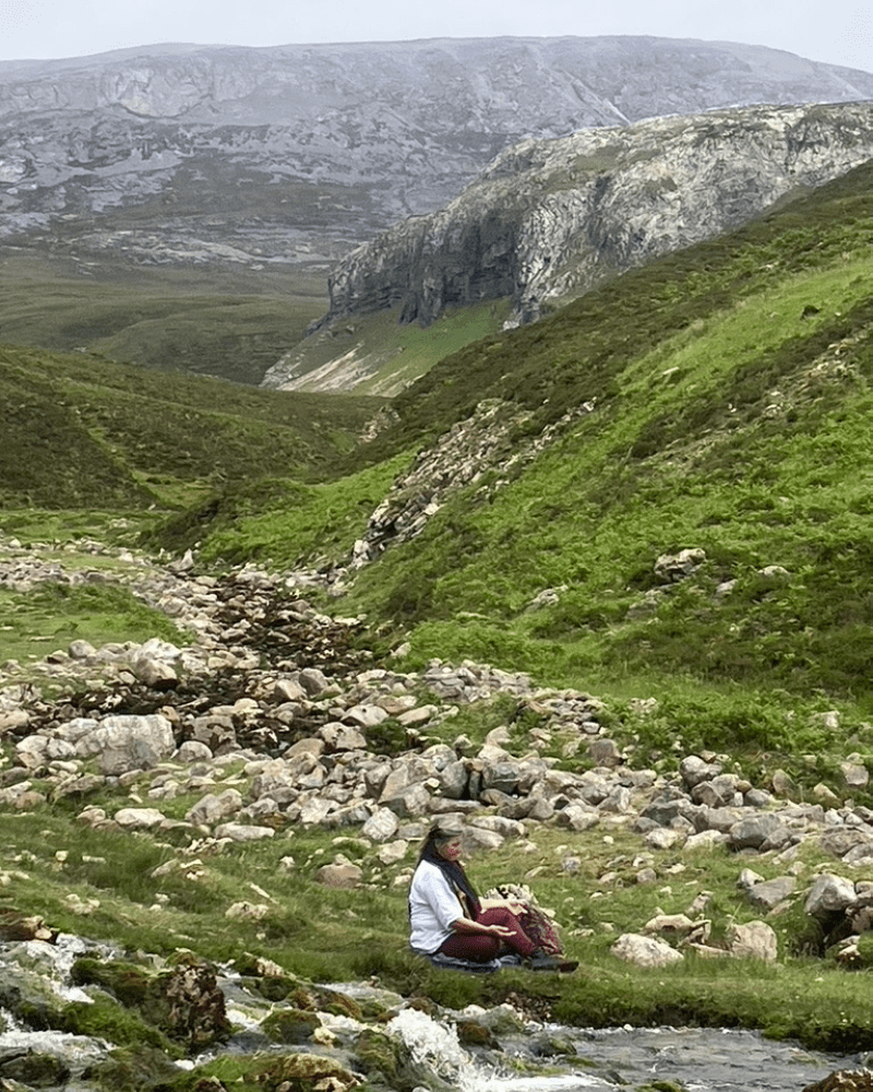 A woman sitrs next to a stream amongst the North West Highlan Mountains, in Scotland